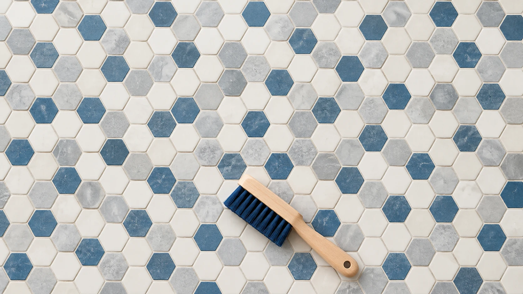 A soft hand brush rests on an intact mosaic floor of white, gray and muted-blue hexagonal tiles.