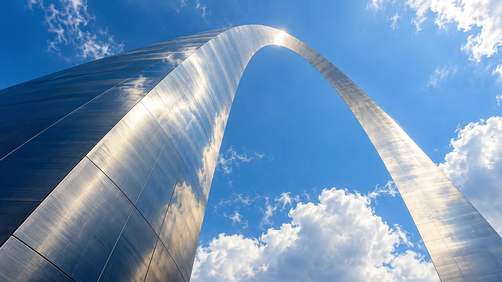 Sunlight reflects off the brushed-metal crown of a sweeping arch against a blue sky.
